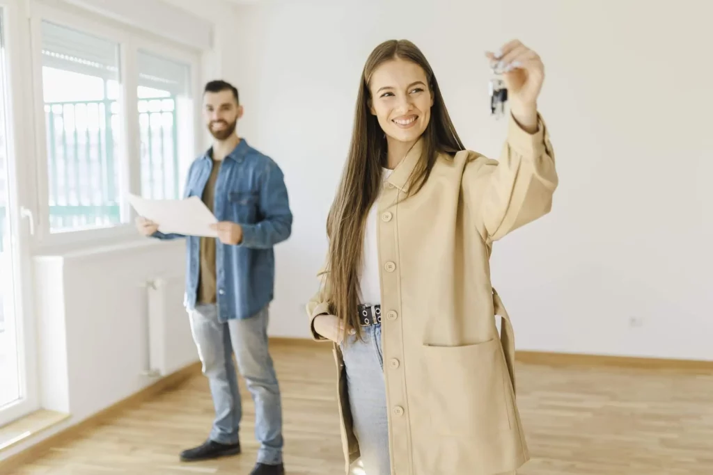 Imagem de um casal composto por um homem e uma mulher em um cômodo vazio para ilustrar matéria sobre o que é cessionário de imóvel. Em primeiro plano, a mulher está segurando a chave da casa. Em segundo plano, o homem está segurando documentos.