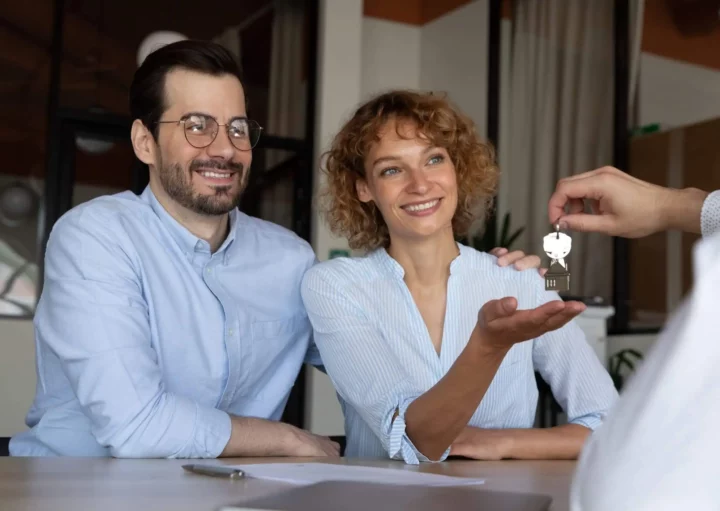 Imagem de um casal sentado à mesa com documentos em cima, recebendo uma chave com chaveiro de casa para ilustrar matéria sobre o que é cessionário de imóvel