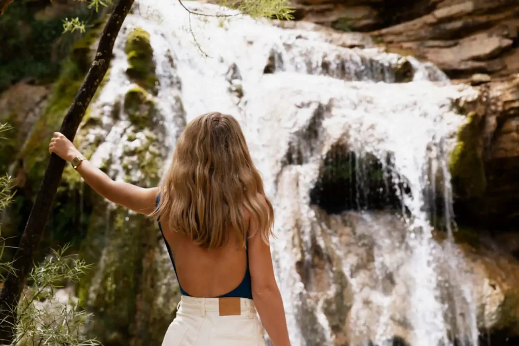 Imagem de uma mulher loira de costas segurando em uma árvore enquanto observa uma cachoeira para ilustrar matéria sobre cachoeiras em SP