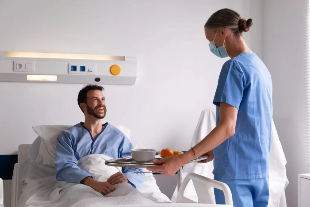 Imagem de um homem sentado na maca de um hospital sorrindo e olhando para uma enfermeira que está em pé na frente dele segurando uma bandeja de café da manhã com algumas frutas dentro do prato para ilustrar matéria sobre cidades com a melhor saúde do Brasil