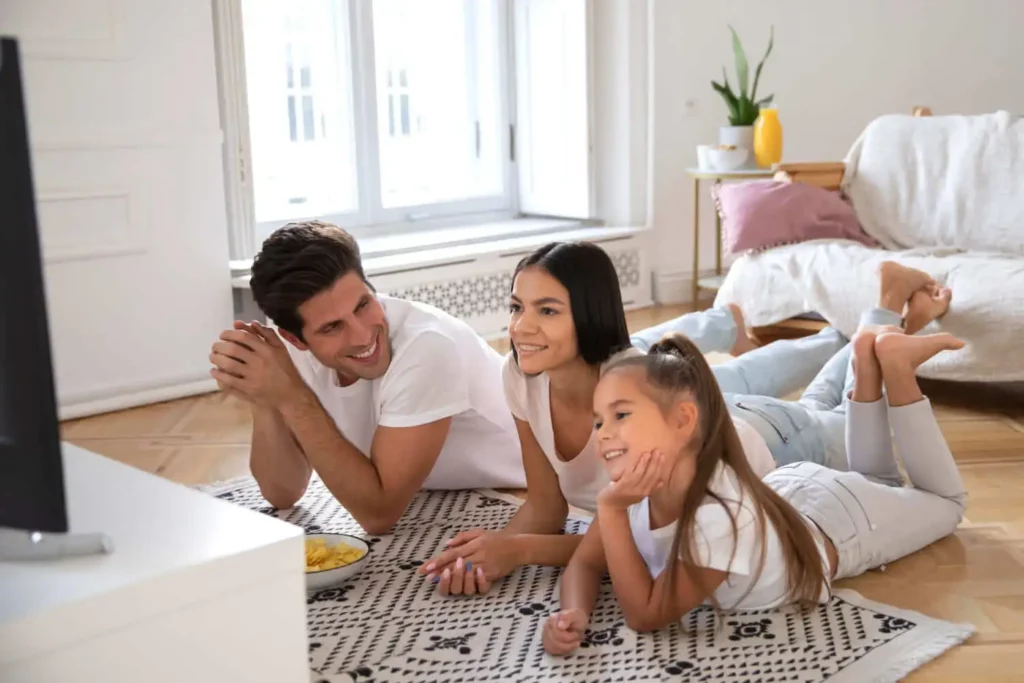 Imagem de uma família composta por um pai, uma mãe e uma criança deitados no chão em frente à tv na sala para ilustrar matéria sobre como saber se o imóvel é novo ou usado