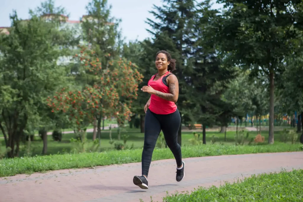 Imagem de uma mulher vestindo camiseta rosa e calça legging preta correndo em uma pista de um parque com vegetação ao redor para ilustrar matéria sobre as desvantagens de morar no centro