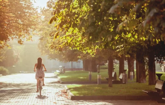magem de uma mulher andando de bicicleta em uma ciclovia em meio às árvores em um parque no fim da tarde para ilustrar matéria sobre parques na Zona Leste de SP