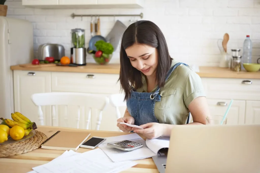 Imagem de uma mulher sentada na mesa da cozinha de uma casa fazendo contas e olhando as informações em um papel para ilustrar matéria sobre como calcular parcelas decrescentes. Em cima da mesa estão frutas, um notebook, calculadora, celular e papéis. Atrás da mulher, está um balcão de cozinha com objetos em cima