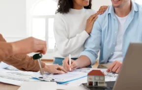 Imagem de um casal composto por um homem e uma mulher assinando um documento que está em cima de uma mesa ao lado de uma casa em tamanho miniatura enquanto outra pessoa entrega uma chave para ilustrar matéria sobre como calcular um financiamento de imóvel