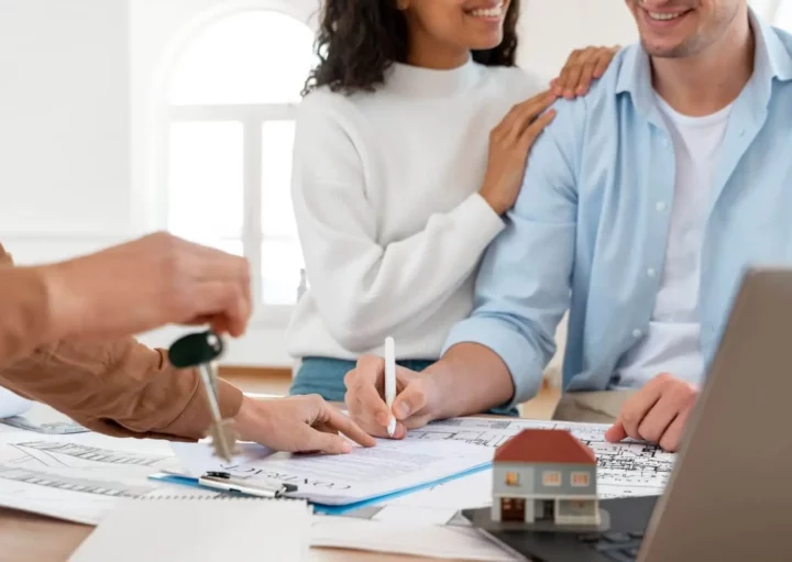 Imagem de um casal composto por um homem e uma mulher assinando um documento que está em cima de uma mesa ao lado de uma casa em tamanho miniatura enquanto outra pessoa entrega uma chave para ilustrar matéria sobre como calcular um financiamento de imóvel