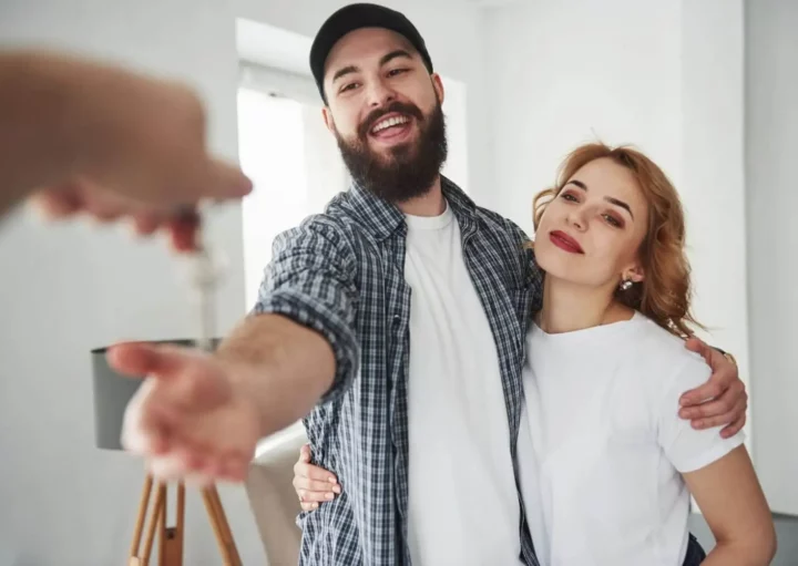 Imagem de um casal formado por um homem e uma mulher se abraçando enquanto recebem a chave de um imóvel de uma terceira pessoa para ilustrar matéria sobre como comprar um apartamento ganhando um salário mínimo