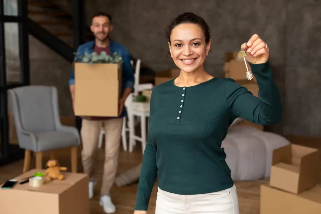 Imagem de um casal composto por um homem e uma mulher em uma casa para ilustrar matéria sobre como conseguir comprar uma casa sendo assalariado. Enquanto a mulher segura a chave de um imóvel, o homem está mais atrás segurando uma caixa de papelão com algumas plantas dentro