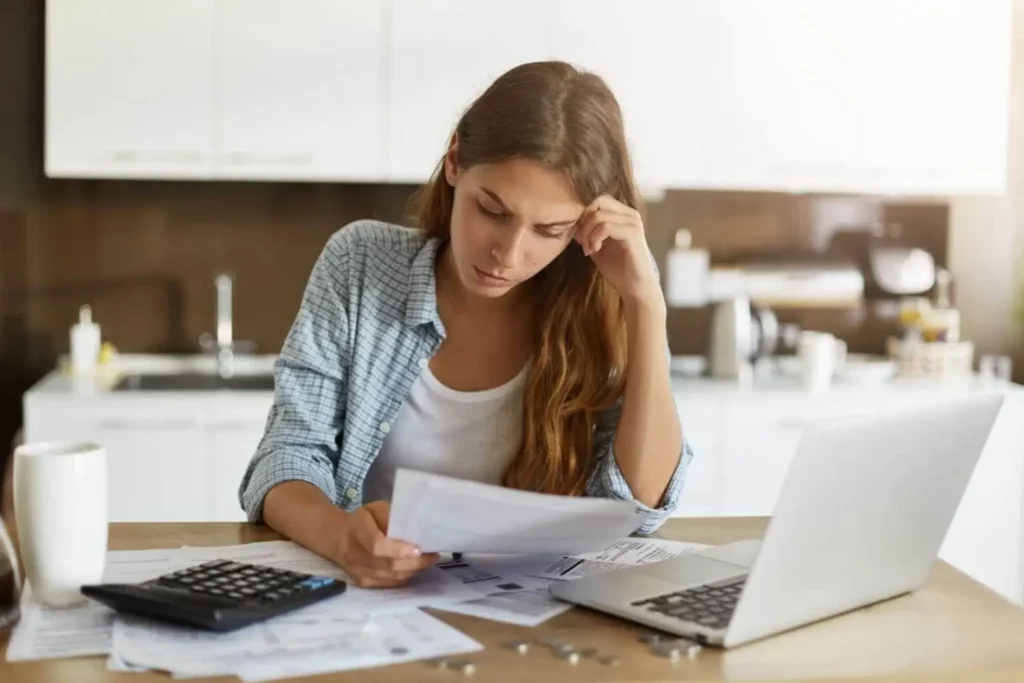 Imagem de uma mulher em frente a um computador que está em cima de uma mesa junto de alguns papéis e uma calculadora para ilustrar matéria sobre como funciona o financiamento condicionado