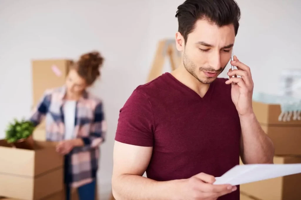 magem de um homem fazendo uma ligação no celular enquanto olha um papel que está em sua mão para ilustrar matéria sobre desistência da compra de imóvel entre particulares. Na mesma imagem, uma mulher ao fundo está tirando objetos de dentro de caixas de papelão