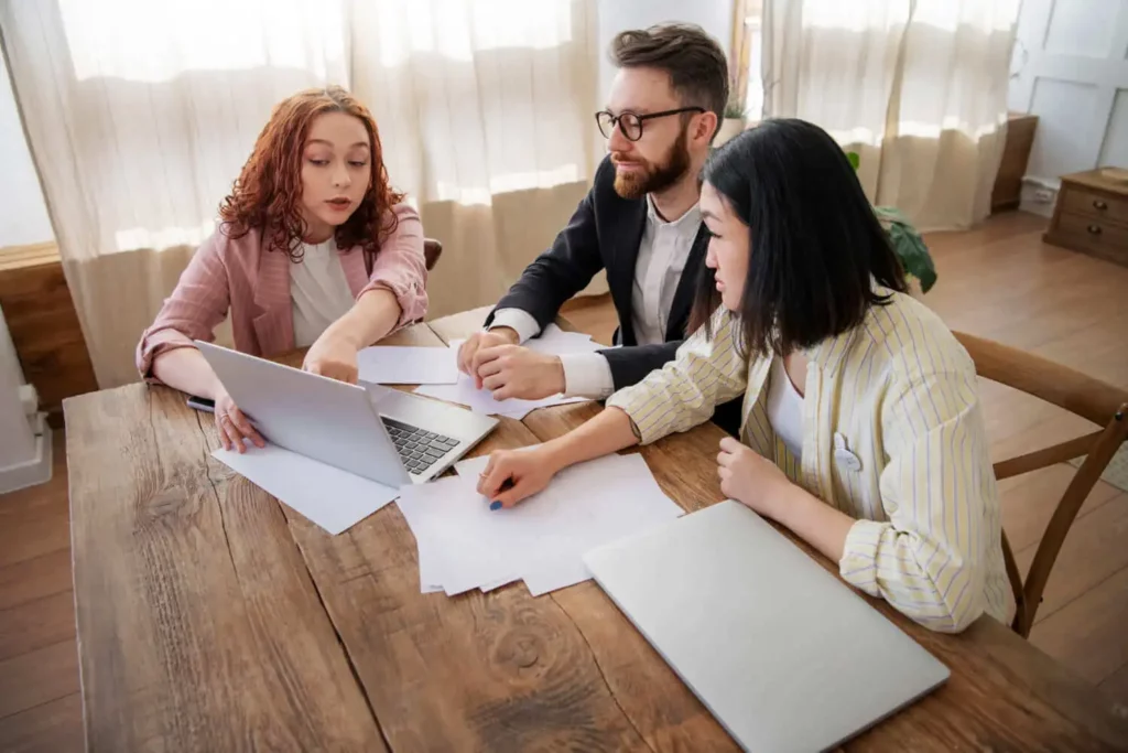 Imagem de um homem e duas mulheres sentados em uma mesa de madeira analisando informações que estão na tela de um notebook, que está ao lado de alguns papéis que estão em cima da mesa, para ilustrar matéria sobre desistência de compra de imóveis entre particulares