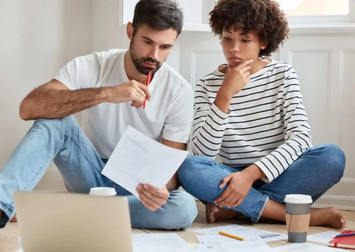 Imagem de um casal composto por um homem e uma mulher sentados no chão de uma casa analisando papéis que estão no chão ao lado de um notebook para ilustrar matéria sobre desistência de compra de imóvel entre particulares