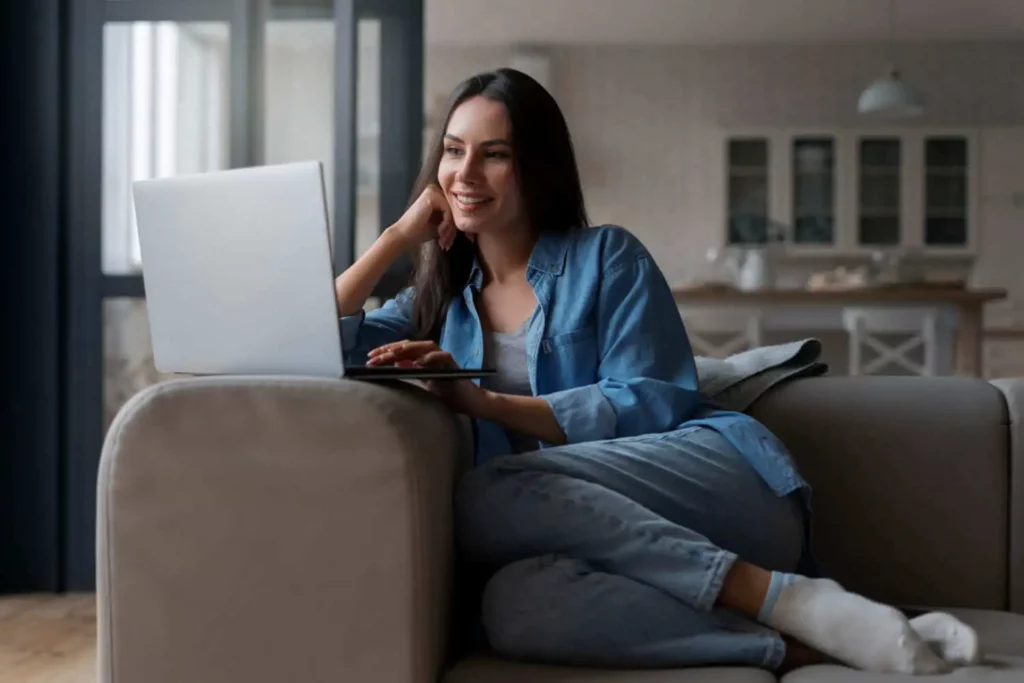 Imagem de uma mulher sentada no sofá de uma casa olhando para o notebook que está em cima do braço do sofá para ilustrar matéria sobre o feirão da Caixa