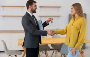 Imagem de um homem e uma mulher apertando as mãos em um escritório para ilustrar matéria sobre feirão de imóveis. A mulher sorri enquanto o homem entrega a chave de um imóvel. Ele tem algumas folhas debaixo do braço. Atrás do homem e da mulher estão uma mesa de madeira, três cadeiras e um armário embutido na parede