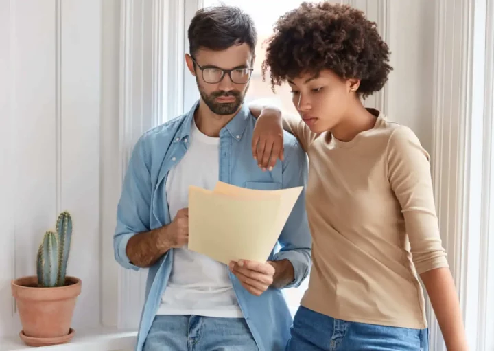 Imagem de um homem e uma mulher um do lado do outro encostados na parede de uma casa olhando para um papel que está na mão do homem para ilustrar matéria que responde a pergunta: imóvel de herança, o cônjuge tem direito?