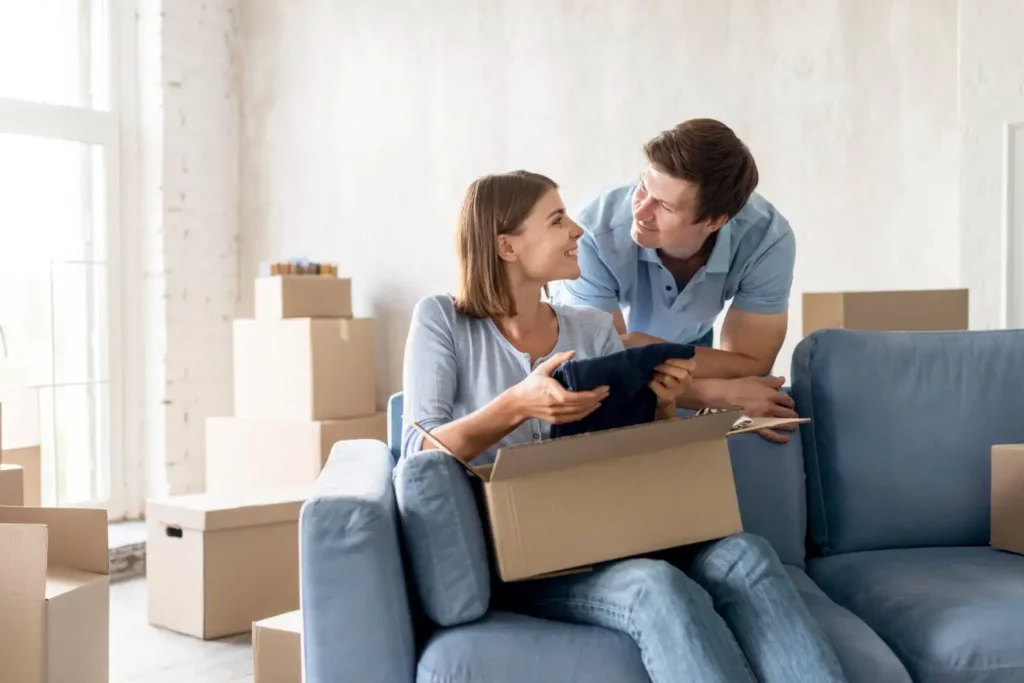 Imagem de um casal composto por um homem e uma mulher em casa para ilustrar matéria sobre se o inadimplente pode financiar imóvel pela Caixa. Enquanto a mulher está sentada no sofá abrindo uma caixa de papelão, o homem está atrás do sofá sorrindo para a mulher.