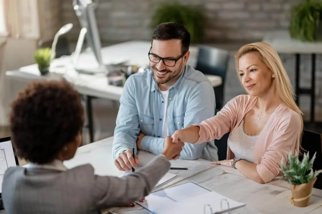 Imagem de uma mulher apertando a mão de uma agente imobiliária enquanto o companheiro observa com um sorriso no rosto para ilustrar matéria sobre o que quer dizer financiamento condicionado