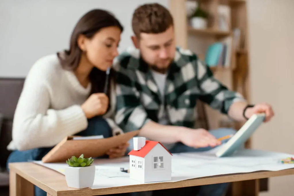 Imagem de um casal sentado no sofá de uma casa analisando informações em uma caderneta para ilustrar matéria sobre parcelas decrescentes ou fixas. Na frente do casal está uma mesinha de centro com alguns objetos em cima, como uma planta suculenta, uma casa em tamanho miniatura e alguns papéis