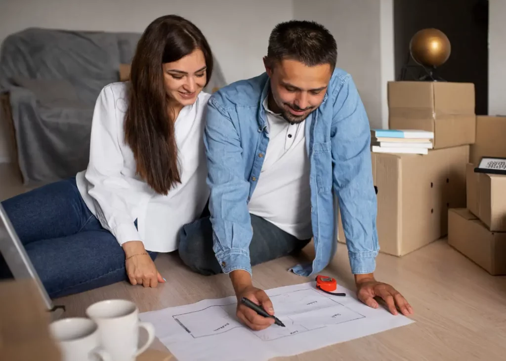 Imagem de um casal formado por um homem e uma mulher sentados no chão de um imóvel com algumas caixas de papelão ao redor e um sofá coberto por uma manta analisando a planta da casa que está no chão para ilustrar matéria sobre o programa habitacional cheque moradia