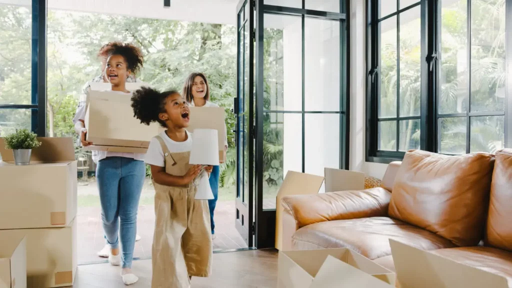 Imagem de uma mulher e duas crianças entrando em um imóvel com algumas caixas de papelão e um abajur na mão para ilustrar matéria sobre quem tem direito ao cheque habitacional