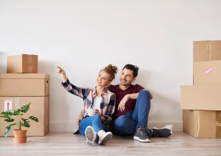 Imagem de um casal formado por um homem e uma mulher sentados no chão de um imóvel ao lado de caixas de papelão e um vaso de planta para ilustrar matéria sobre se quem tem restrição no nome pode financiar um imóvel pela Caixa