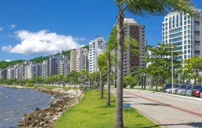 Imagem de prédios, árvores, gramado e pista para caminhada na beira do mar em um bairro da capital de Santa Catarina para ilustrar matéria sobre bairros bons e baratos para morar em Florianópolis