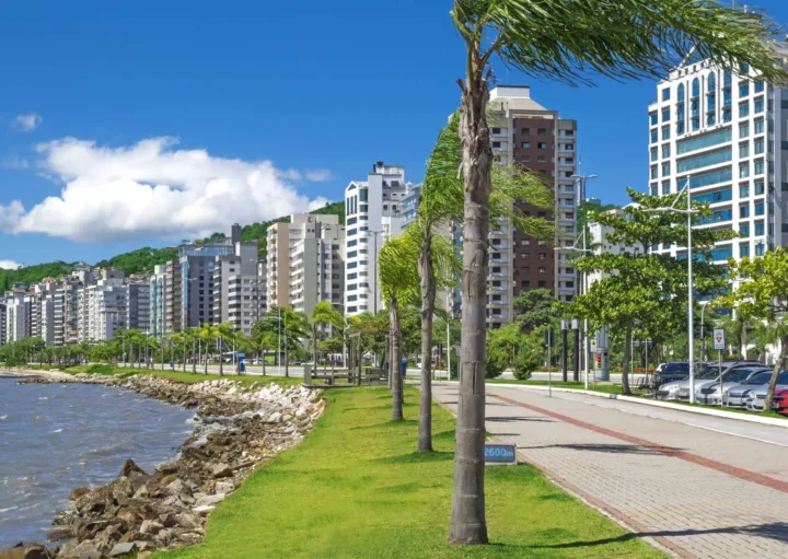 Imagem de prédios, árvores, gramado e pista para caminhada na beira do mar em um bairro da capital de Santa Catarina para ilustrar matéria sobre bairros bons e baratos para morar em Florianópolis