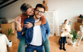 Imagem de uma mulher sentada nas costas de um homem que está em pé sorrindo enquanto o abraça para ilustrar matéria sobre como ganhar dinheiro com imóveis. Eles estão em uma casa com algumas caixas de papelão e uma criança segurando um urso de pelúcia