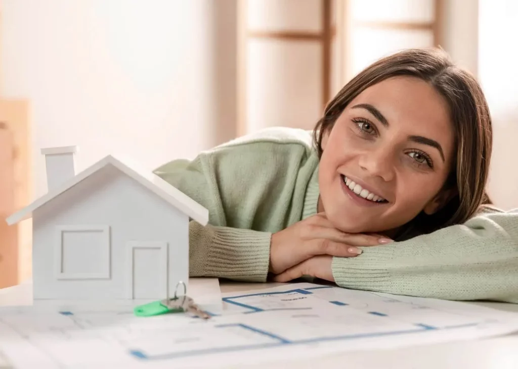 Imagem de uma mulher encostada em cima de uma mesa onde estão as chaves de um imóvel, a planta do imóvel e uma casa de madeira na cor branca em tamanho miniatura para ilustrar matéria sobre como investir em imóveis com ações de construtoras com pouco dinheiro