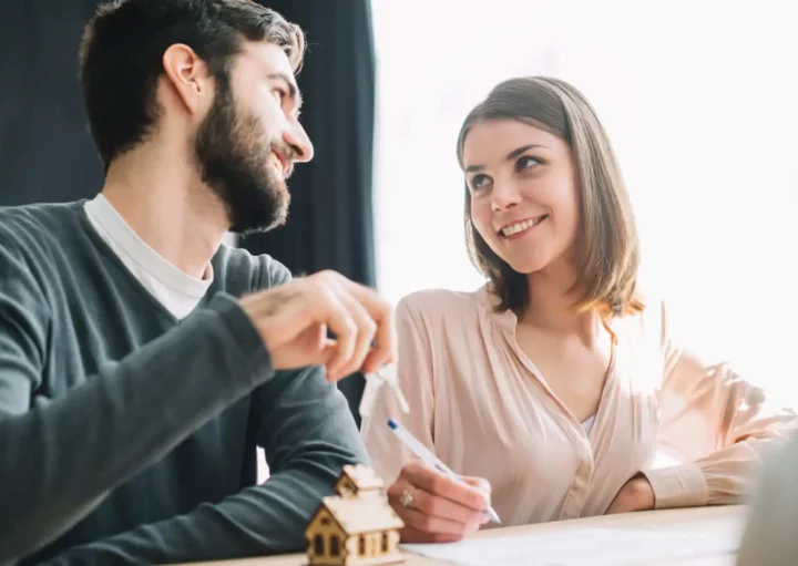 Imagem de um casal formado por um homem e uma mulher se olhando enquanto assinam um documento que está em cima de uma mesa ao lado de uma casa de maneira em tamanho miniatura para ilustrar matéria sobre como investir em imóveis com pouco dinheiro