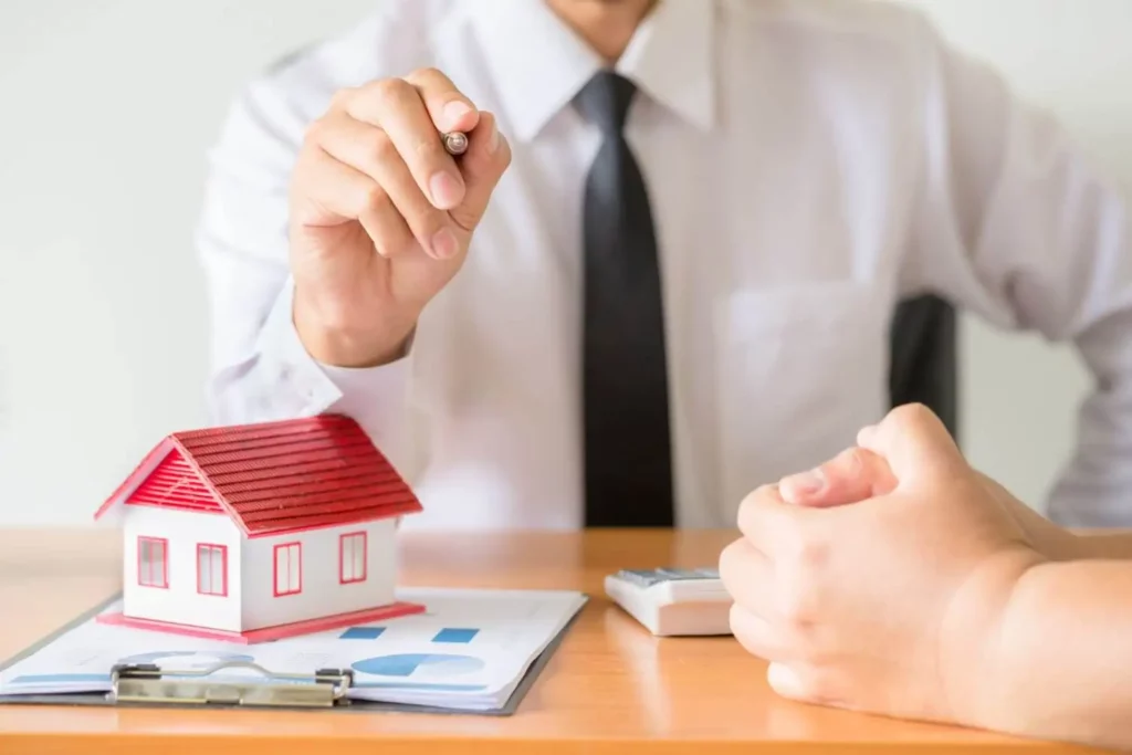 Imagem de um corretor imobiliário usando camisa social e gravata preta em frente a outra pessoa apontando com uma caneta enquanto uma prancheta e uma casa de madeira em tamanho miniatura estão em cima de uma mesa para ilustrar matéria sobre como investir em imóveis tendo pouco dinheiro