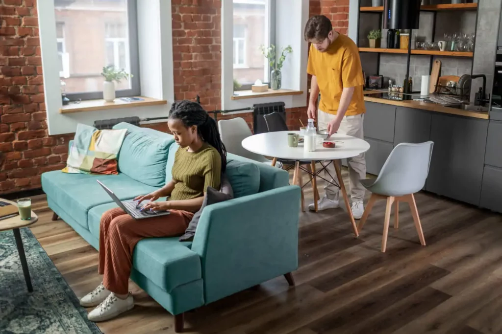 Imagem de um homem e uma mulher em uma sala e cozinha integrada de uma kitnet para ilustrar matéria sobre como planejar a construção de kitnet para alugar. Enquanto a mulher está sentada no sofá da sala digitando no computador, o homem está em pé em frente a uma mesa branca na cozinha se servindo em algumas tigelas