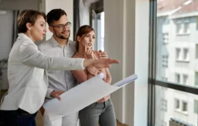 Imagem de uma agente imobiliária com uma planta na mão explicando sobre o imóvel para um casal formado por um homem e mulher para ilustrar matéria sobre garantia de imóvel novo