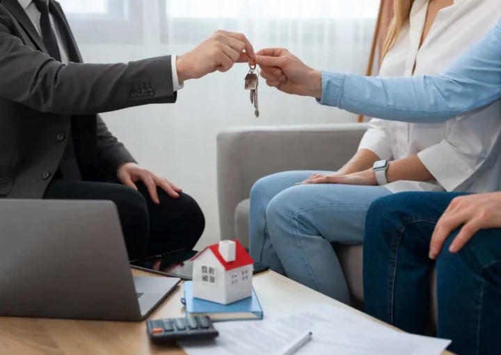 Imagem de um agente imobiliário entregando a chave para um casal próximo a uma mesa cheia de documentos, um computador para e um modelo de casa para ilustrar matéria sobre quanto tempo leva para comprar um imóvel