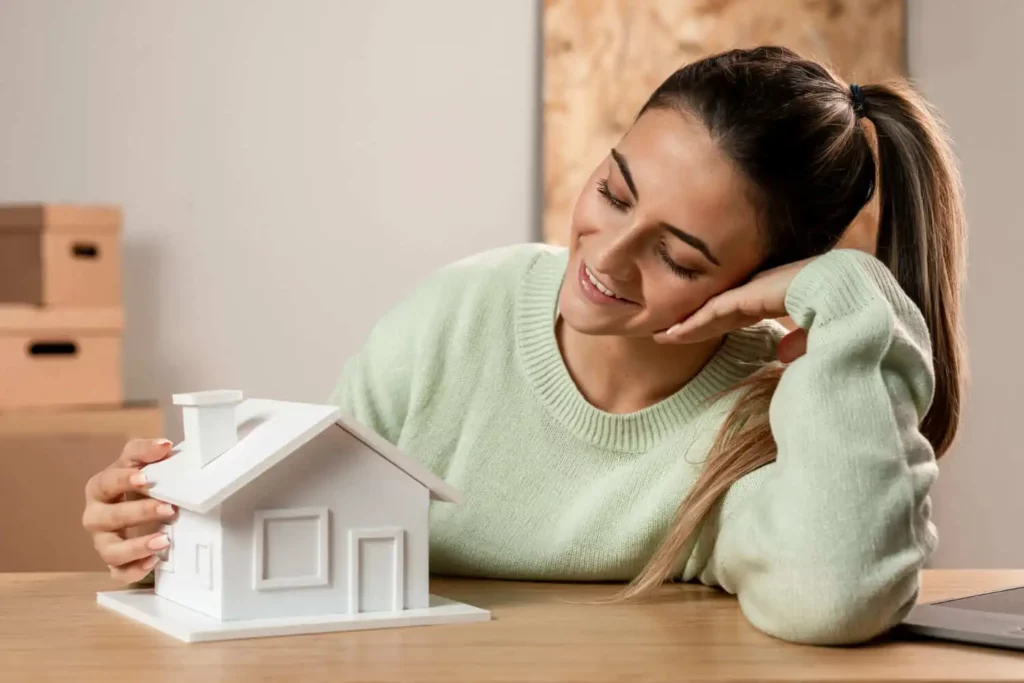 Imagem de uma mulher apoiando a cabeça com o cotovelo em cima de uma mesa enquanto olha para uma casa de madeira em tamanho miniatura que está em cima da mesa para ilustrar matéria sobre o tempo médio para vender imóvel