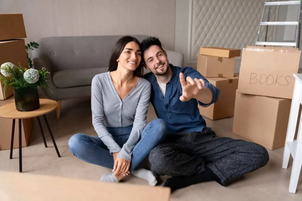 Imagem de um casal formado por um homem e mulher sentados no chão da sala de uma casa cercados por um sofá, uma mesa de centro e algumas caixas de papelão para ilustrar matéria se vale a pena não vender imóvel