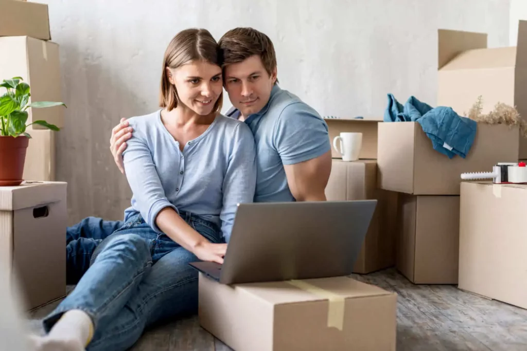 Imagem de um casal composto por um homem e uma mulher sentados no chão de um imóvel ao redor de várias caixas de papelão empilhadas olhando para a tela de um notebook que está em cima de uma das caixas para ilustrar o tempo médio para vender uma casa